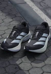 Chaussures de course Adidas Adizero noir et blanc posées sur un pavé en pierre hexagonal près du bord de la route.