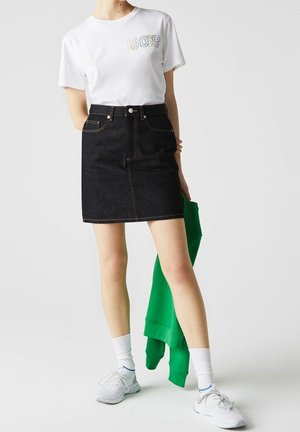 Femme portant un t-shirt blanc à motif graphique et une jupe en jean noire, tenant une veste verte, avec des chaussettes blanches et des baskets sur un fond uni.