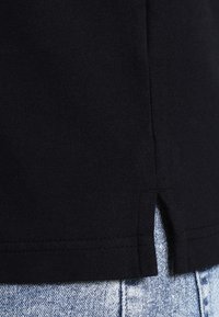 Černé polokošile z látky s texturovaným tkaním, s bočním rozparkem na lemu, nošené přes světle modré džíny.