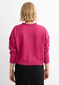 Suéter de punto en rosa brillante con cuello redondo y hombros caídos, con puños y dobladillo de canalé, llevado sobre una falda negra.