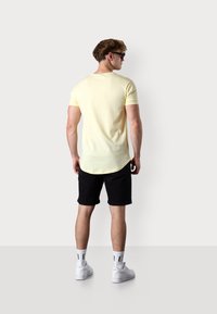 Yellow short-sleeve shirt, black shorts, white sneakers, and white socks. Rear view showcasing the shirt’s curved hem and casual fit.