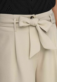 Beiga byxor med ett brett tygbälte knutet i en sløyfe. Har en slät struktur, rak passform och en knappstängning i midjan.