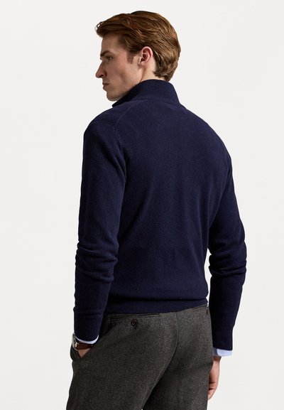 Marineblauwe wolmix trui met een hoge kraag, getailleerde silhouet en ribgebreide manchetten. Gecombineerd met grijze textuurbroek en zichtbare blauwe overhemdmanchetten.