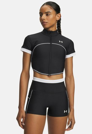 Vrouw draagt een zwarte, korte, met rits sluitende sporttop met witte bies en bijpassende zwarte shorts met hoge taille, wit logo en bies, staand met ontspannen armen.