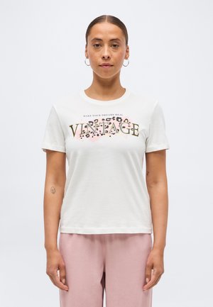 Naine kannab valget t-särki, millel on tekst „VINTAGE“ ja leopardimustriga disain, ning helepunaseid pükse, seistes ühtlase tausta ees.