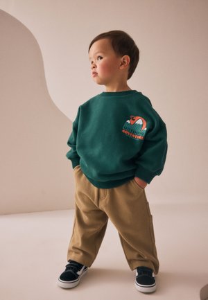 Toddler wearing a green sweatshirt with "Outdoor Adventure" graphic, tan pants, and black sneakers, standing with hands in pockets, looking right.