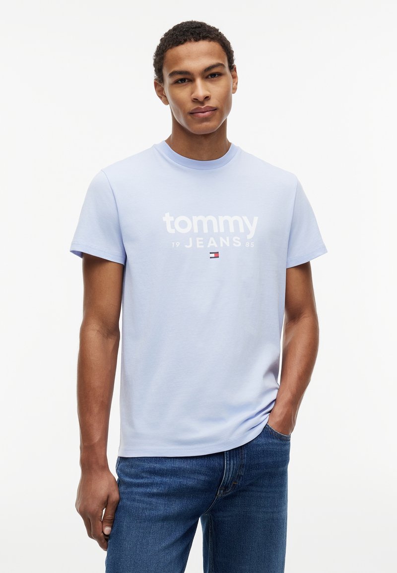 Jeune homme debout, une main dans la poche, portant un t-shirt bleu clair « Tommy Jeans » et un jean bleu sur fond blanc.