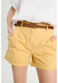 Shorts en coton jaunes avec un ourlet roulé, des plis et des poches sur les côtés, complétés par une ceinture en cuir tressé brun.