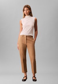 Sieviete stāv, tērpusies baltā bezrokātopā, gaiši brūnās augsta jostasvieta pielāgotās biksēs un melnās dzīvoklās kurpēs, viena roka kabatā.