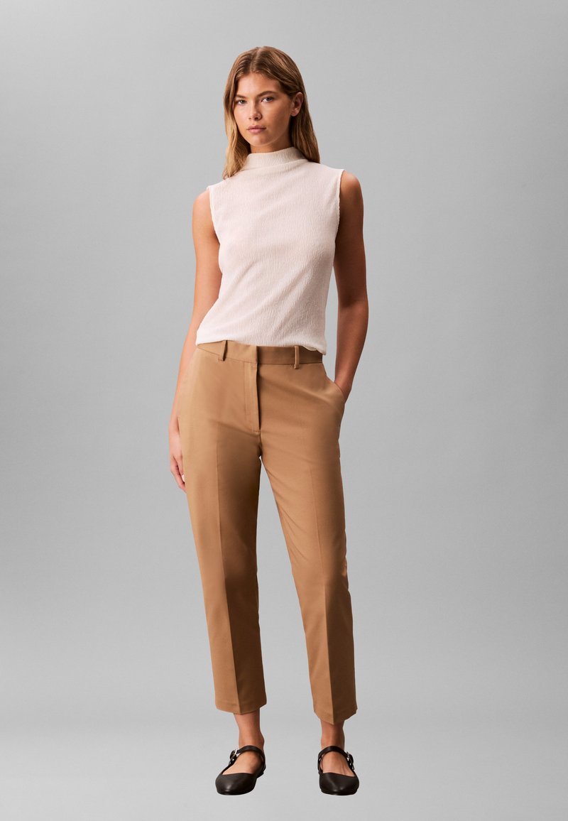 Sieviete stāv, tērpusies baltā bezrokātopā, gaiši brūnās augsta jostasvieta pielāgotās biksēs un melnās dzīvoklās kurpēs, viena roka kabatā.