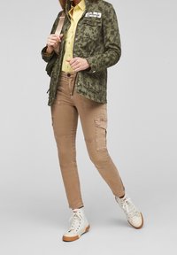 Persoon in beige cargobroek, witte hoge sneakers, gele shirt en groene camouflagejas met "Dreamers" patch, met een rugzak.
