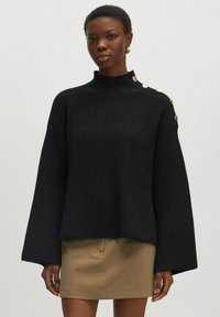 Schwarzer Strickpullover mit hohem Kragen, weiten Ärmeln und goldenen Knopfdetails an der Schulter, kombiniert mit einem beigen Rock.