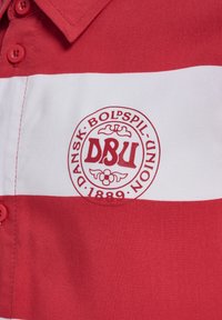 Maglietta a righe rosse e bianche con colletto, caratterizzata da un logo rotondo che riporta la scritta "DBU" e "DANSK BOLSPIL-UNION 1889." Tessuto testurizzato.