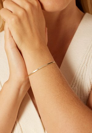 Gouden metalen bangle om een pols, slank en glad met een glanzende afwerking. De huidskleur is licht, met een gestructureerde gebreide stof op de achtergrond.