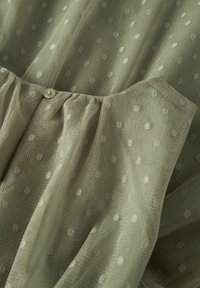 Tissu texturé vert clair avec des pois blancs, présentant des détails froncés et une fermeture à bouton. Composition douce et semi-transparente.