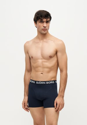Homme musclé aux cheveux foncés portant un boxer Björn Borg bleu marine, debout devant un fond clair uni, regardant devant lui.