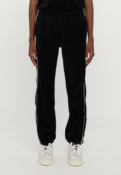 Joggers en velours noir avec des rayures blanches sur les côtés, une taille élastique et des poignets. Présentent une texture lisse et une coupe décontractée. Associés à des baskets blanches.