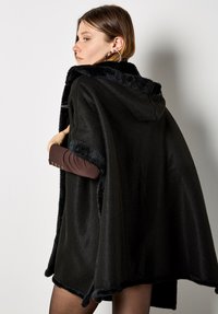 Schwarzer Kapuzenponcho aus strukturiertem Stoff mit flauschigem Besatz, offenen Seiten und einem Saum, der bis zur Mitte des Oberschenkels reicht.
