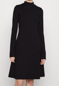 Robe noire à manches longues avec un col haut et un motif de texte répétitif, faite de tissu lisse avec un corsage ajusté et une jupe évasée.