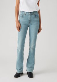 Jeans de mezclilla azul claro con un diseño de piernas acampanadas, que cuentan con bolsillos frontales y cierre de cremallera. Llevados con una camiseta blanca y botas negras.