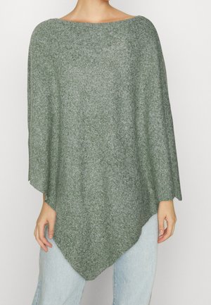 Mujer con un poncho verde suelto y texturizado con dobladillo asimétrico sobre unos vaqueros azul claro, de pie frente a un fondo blanco liso.
