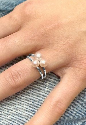 Main portant une bague argentée à double bande ornée de trois perles blanches rondes, posée sur un tissu en denim bleu.