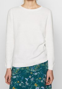 Biały sweter dzianinowy z okrągłym dekoltem i długimi rękawami. Posiada teksturowany wzór oraz ściągacz w pasie. W połączeniu z spódnicą w kwiatowy wzór.