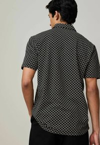 Camicia a maniche corte in tessuto nero con un motivo geometrico bianco. Presenta un colletto e una vestibilità rilassata, con dettagli di cucitura visibili.