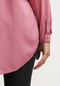 Chemise rose pâle à coupe décontractée, manches longues, poignets boutonnés et ourlet incurvé. Texture lisse, tissu qui tombe doucement.