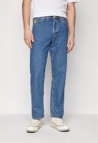 Man i raka blå jeans med en gul leende ansikte-patch på fickan och beige plattformssneakers.