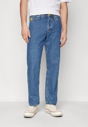 Mann trägt gerade geschnittene blaue Jeans mit einem gelben Smiley-Patch auf der Tasche und beigefarbenen Plattform-Sneakern.