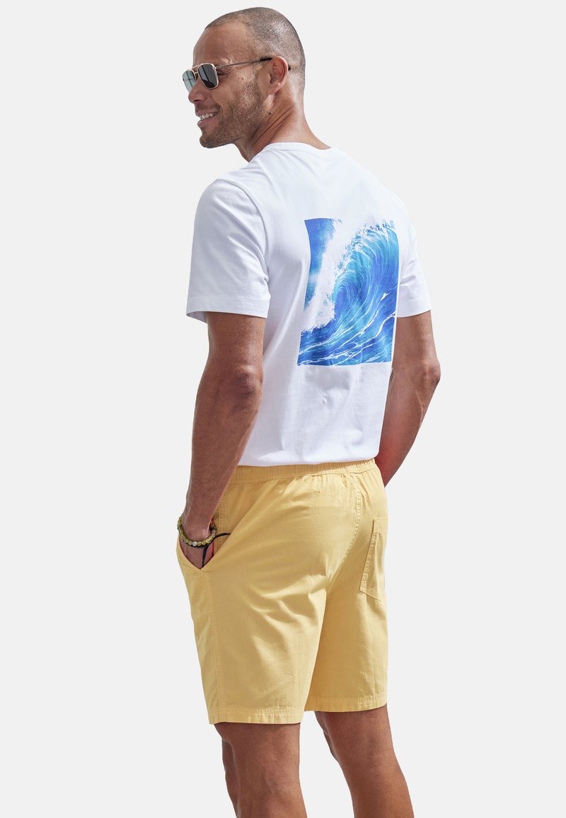 Weißes T-Shirt mit blauer Wellengrafik auf der Rückseite, kombiniert mit gelben Shorts. Hat einen lässigen Schnitt und Taschen in den Shorts.