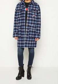 Cappotto a quadri blu navy con accenti azzurri, caratterizzato da grandi bottoni, due tasche frontali e una vestibilità rilassata. Indossato con jeans skinny e stivali neri.