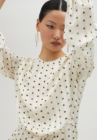 Blusa crema con lunares negros y rayas verticales. Presenta mangas amplias y un escote redondeado. Pendientes geométricos dorados.