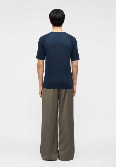 Άτομο που φαίνεται από πίσω, φοράει μπλε σκούρο κοντομάνικο πουκάμισο και καφέ φαρδιά, άνετα παντελόνια με φαρδύ πόδι, με φόντο ένα απλό υπόβαθρο.