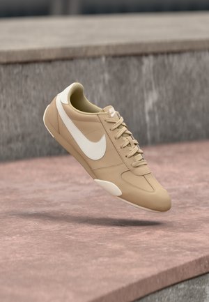 Pantof sport Nike bej cu logo alb și șireturi albe, în aer, deasupra unei suprafețe de beton, cu trepte în fundal.