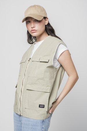 Khaki sleeveless vest with multiple front pockets and a front zipper, paired with a beige baseball cap, worn over a white shirt.