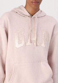Sudadera rosa clara hecha de una tela suave, con un gran logo de "GAP" en acabado satinado, bolsillo tipo canguro y capucha con cordón.