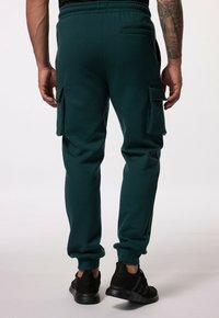 Dunkelgrüne Jogginghose mit Kordelzugbund, seitlichen Taschen und Cargo-Taschen im Cargo-Stil an den Oberschenkeln. Weicher Stoff und schmal zulaufende Bündchen.