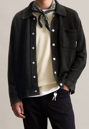 Person trägt schwarze, geknöpfte Jacke über beigem Pullover mit einem Schlüsselanhänger, der aus der Hosentasche der schwarzen Hose hängt, und einem gemusterten Halstuch.