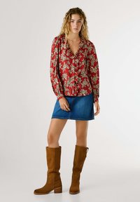 Blouse florale rouge avec un col froncé, portée avec une jupe en jean et des bottes en daim marron jusqu'au genou. Présente des manches longues et une coupe décontractée.