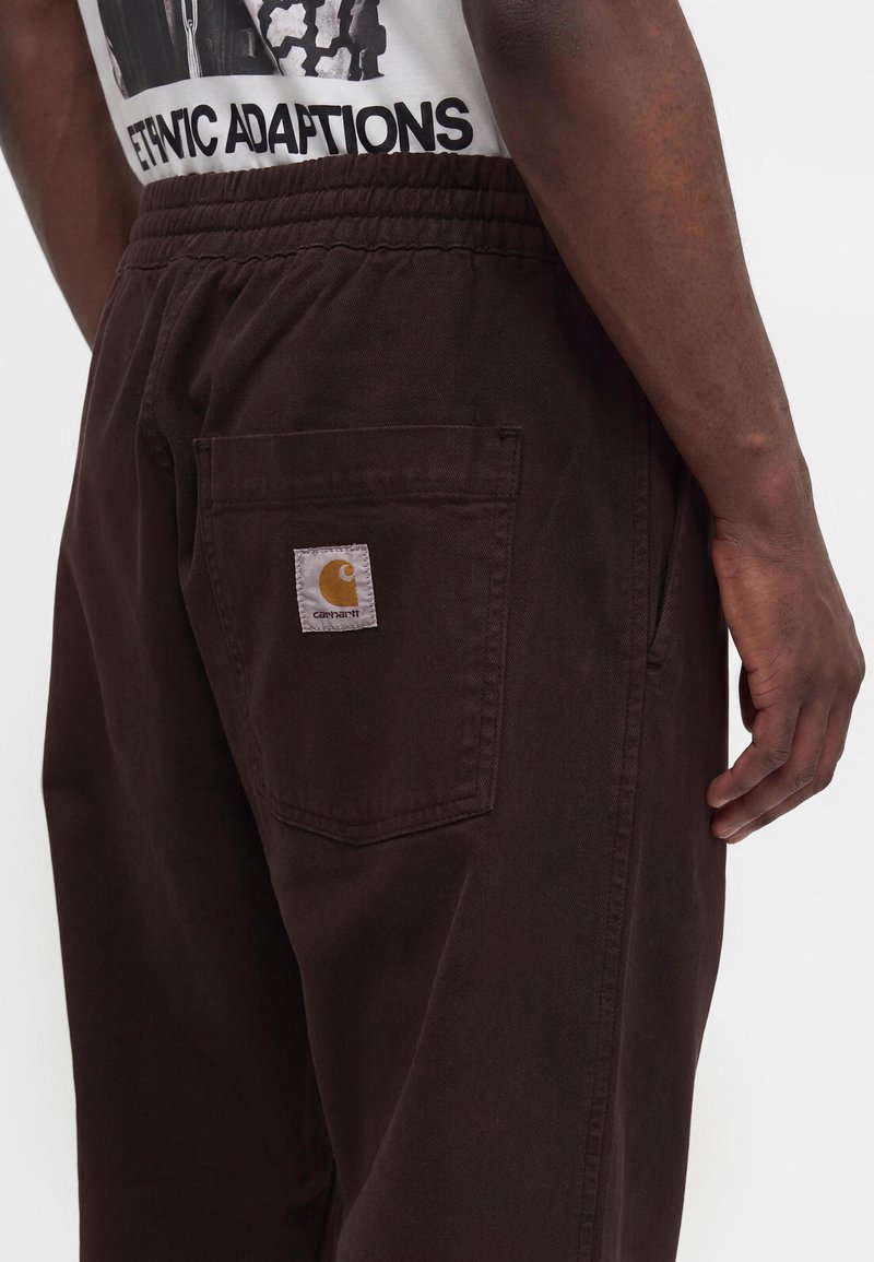 Uomo che indossa pantaloni Carhartt marrone scuro con tasca a toppa posteriore e una t-shirt bianca con stampa grafica che recita "ETHNIC ADAPTATIONS."