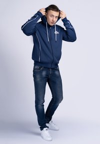 Marineblå zip-hoodie med hvide detaljer, forlommer og justerbare snore. Kombineret med tætsiddende mørke jeans og hvide sneakers.