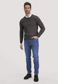 Dunkelgrauer Strickpullover mit rundem Halsausschnitt, kombiniert mit hellblauen Jeans und dunkelbraunen Schuhen. Das Model steht mit den Händen in den Taschen und lächelt.