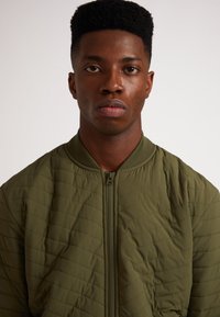 Giacca bomber trapuntata verde oliva con zip frontale e colletto a coste, caratterizzata da cuciture orizzontali e una texture morbida.