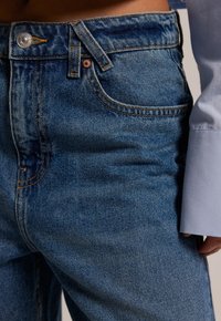 Blue denim jeans featuring a mid-rise waist, light fading, classic five-pocket design, and contrasting orange stitching details.