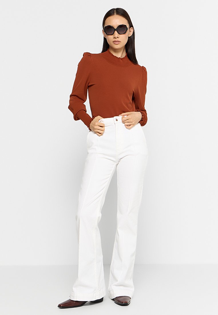 Braunes Langarmoberteil mit Rüschärmeln, kombiniert mit hochtaillierten weißen Schlagjeans und schwarzen Sonnenbrillen. Glatte Texturen und figurbetontes Design.