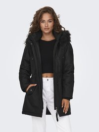 Black parka jacket featuring a faux fur-lined hood, zip and button closure, and two front pockets. Worn with a black knit top and white pants.
