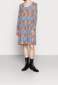 Vestido de manga larga con un patrón circular en azul y naranja, con escote redondeado y falda plisada; combinado con botas negras hasta el tobillo.