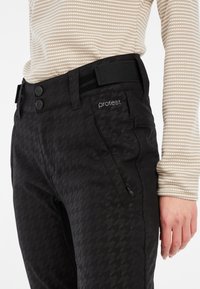 Schwarze Schneehose mit Hahnentrittmuster, ausgestattet mit einem Knopfverschluss, Gürtelschlaufen und seitlichen Reißverschlusstaschen. Der Stoff wirkt strukturiert.